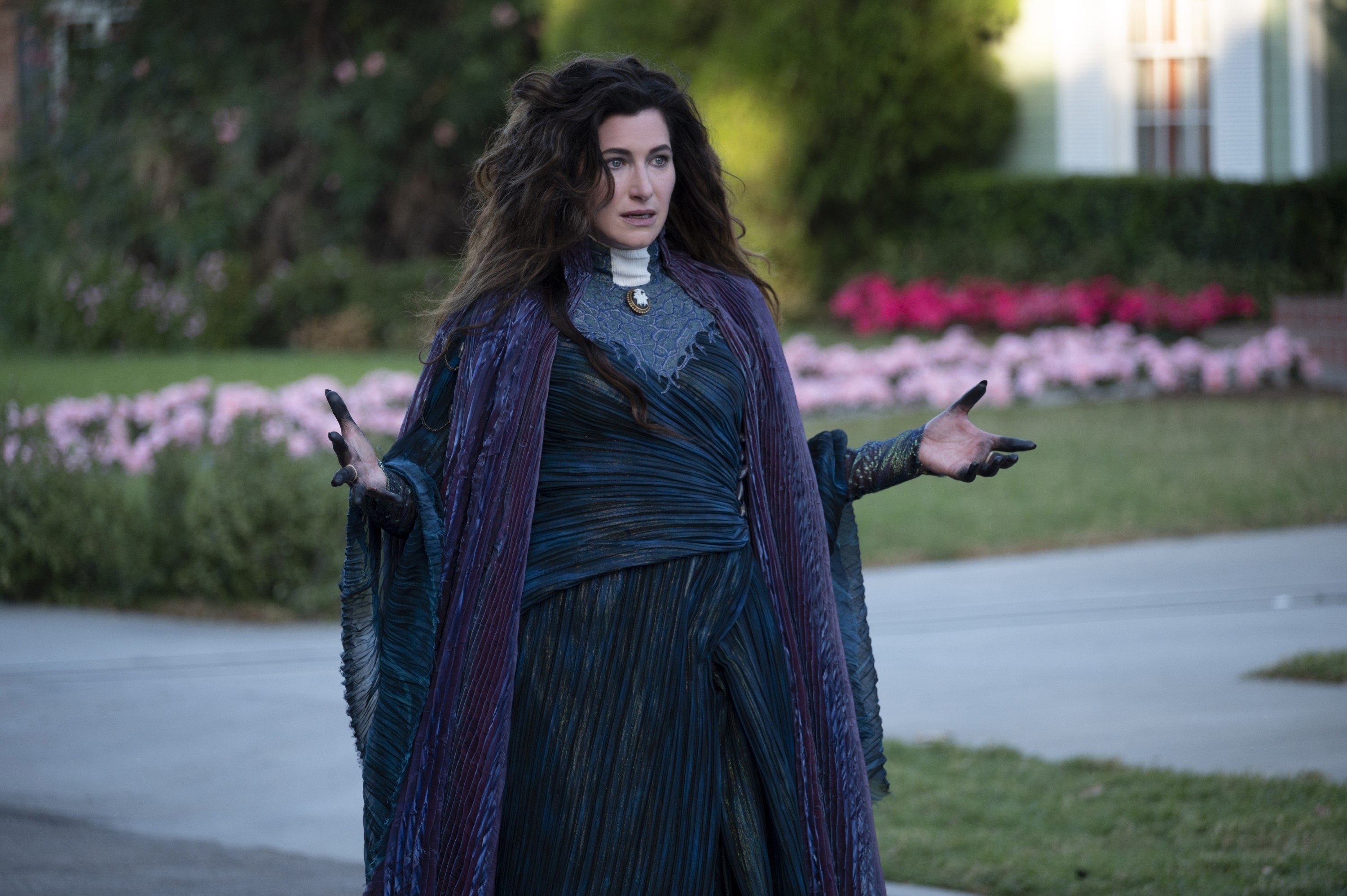 Kathryn Hahn as Agatha wearing a flowing dress and cape, standing outdoors with her hands outturned