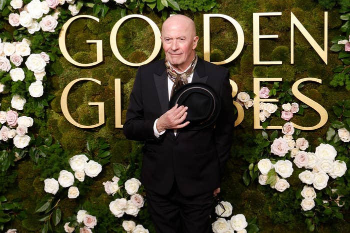 Person in formal suit holding a hat, posing at a Golden Globes event backdrop with floral decorations