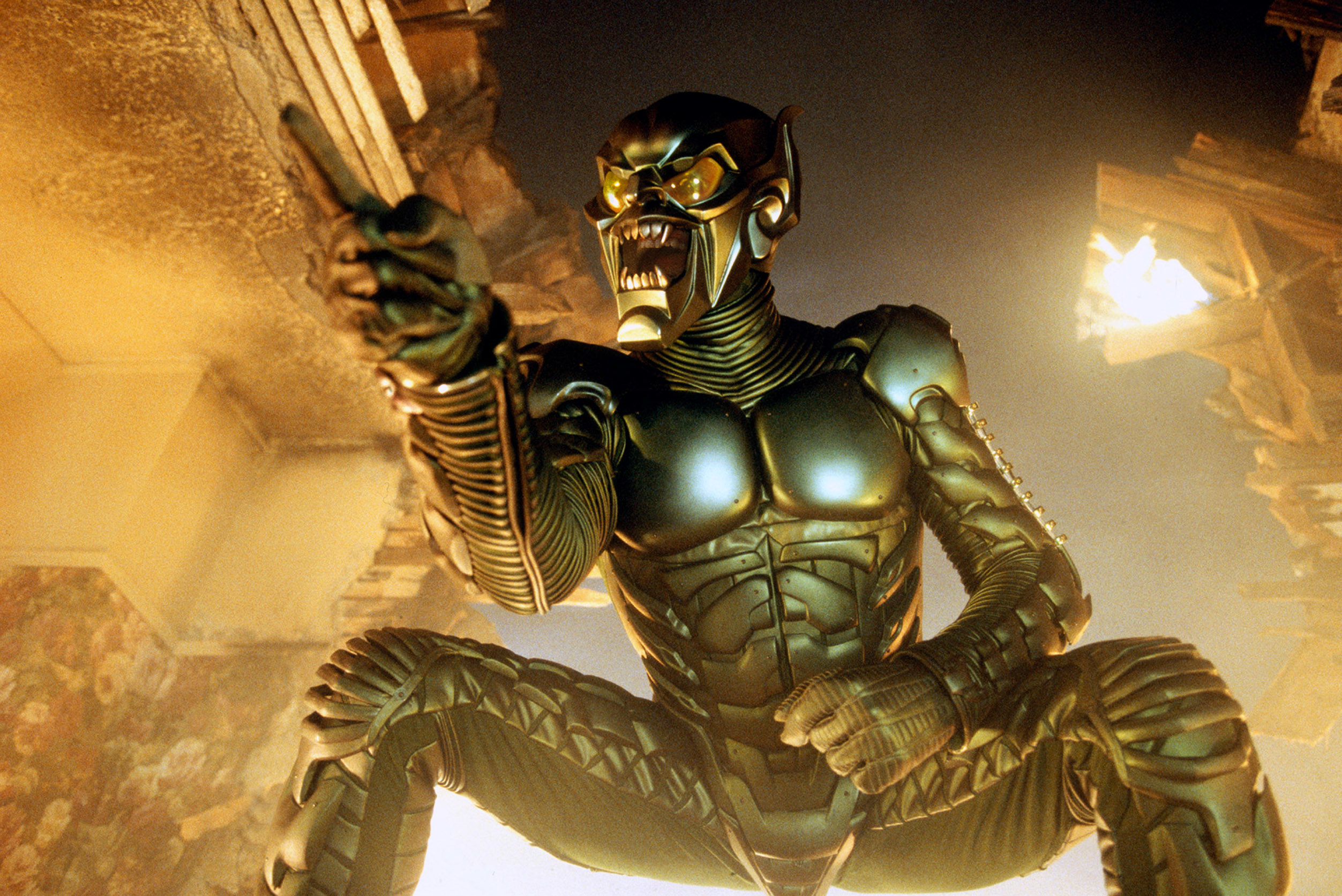 Willem Dafoe as Green Goblin in a metallic armored suit and helmet poses dramatically in a squat and pointing
