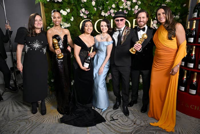 Group of people at an award event, holding trophies. Some are wearing formal gowns and suits, posing in front of a floral backdrop
