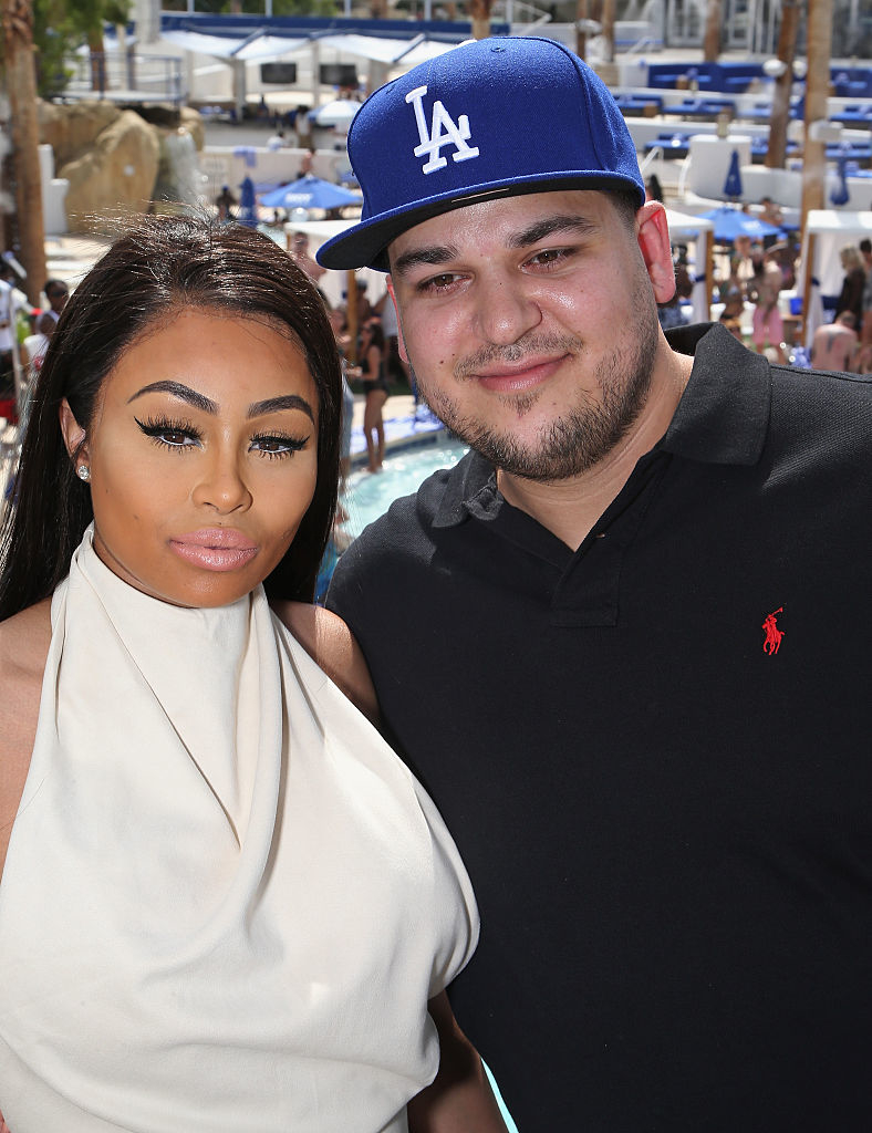 Two people posing at a pool party. One is wearing a baseball cap and polo shirt, and the other is in a draped halter top. Crowd in the background