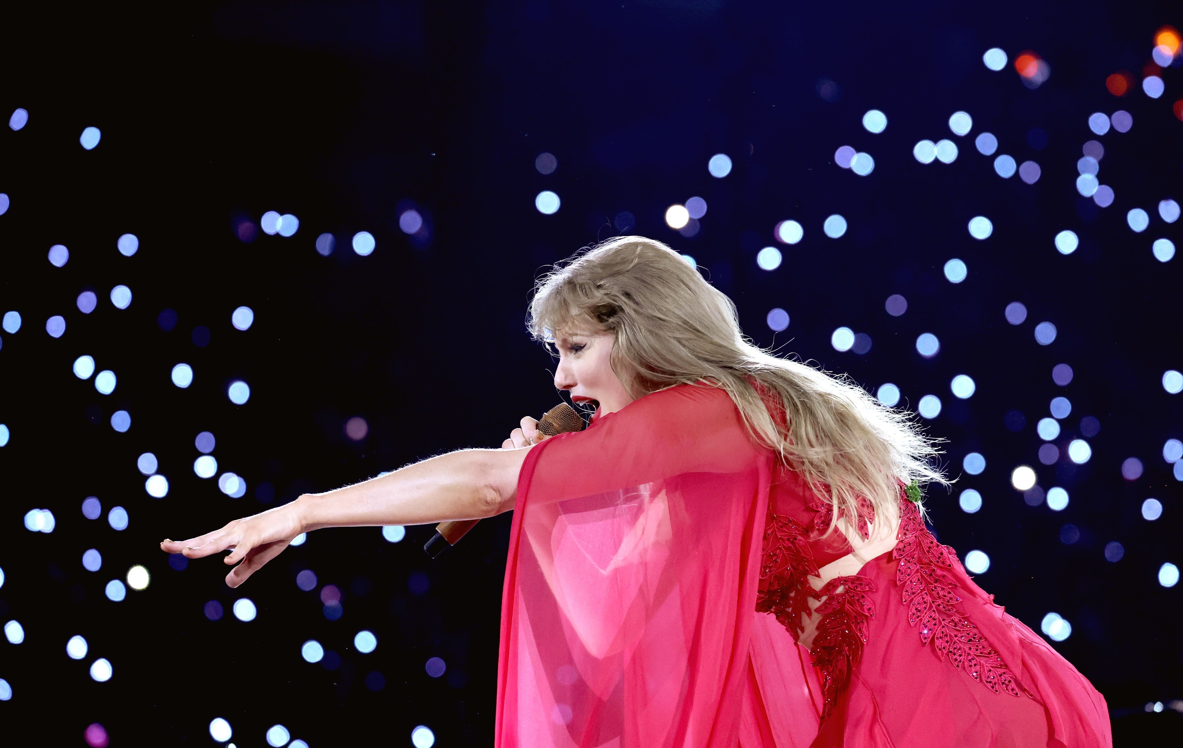 Taylor Swift performing on stage in an elegant, flowing outfit, reaching out to the audience amidst a backdrop of sparkling lights