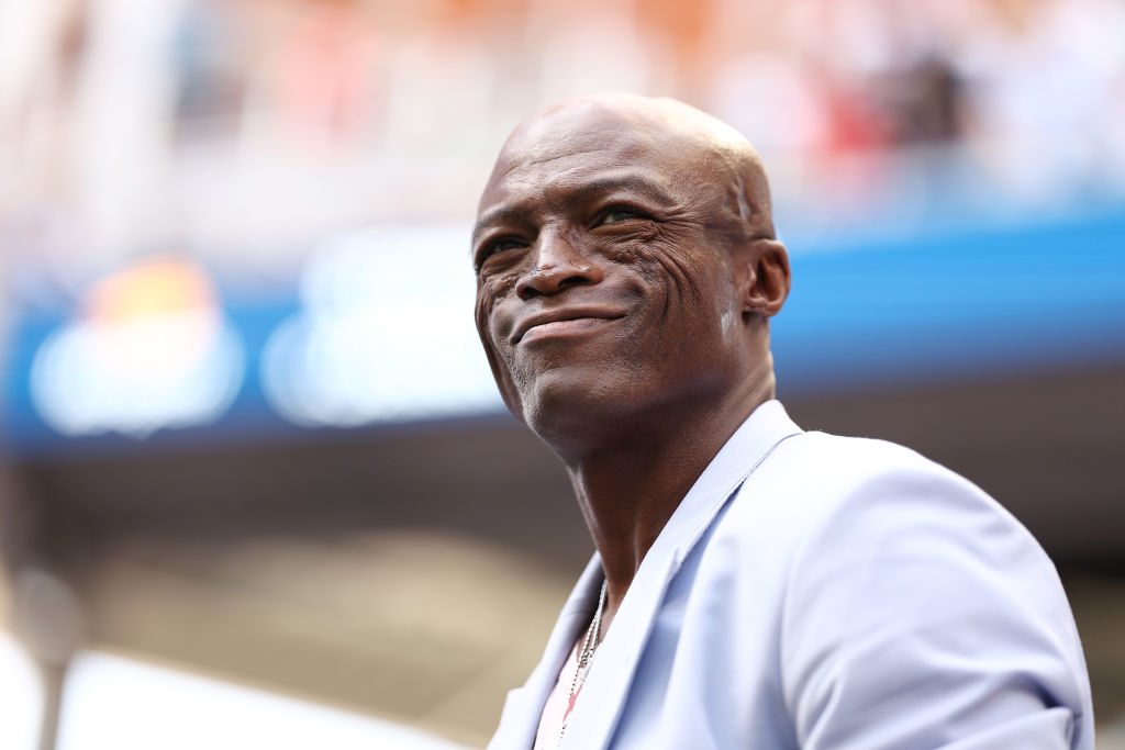 Person smiling outdoors, wearing a light blazer, looking content. Blurred background suggests an open-air event