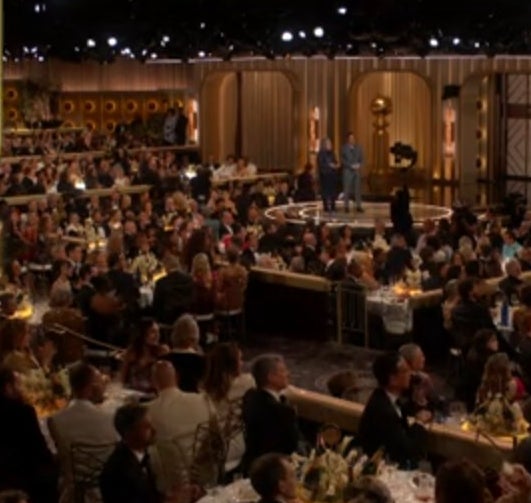 An audience watches an award ceremony. An inset shows Hannah Einbinder in a one-shoulder dress accepting an award with a speech