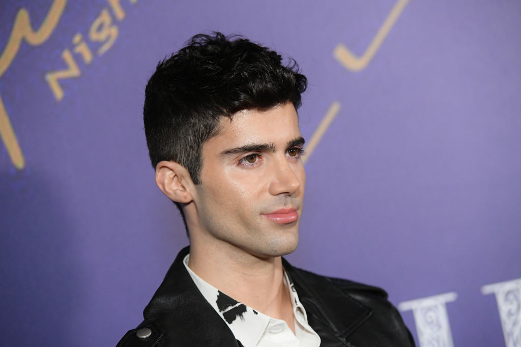 Person in a black jacket over a white shirt at an event, standing against a purple backdrop with partially visible text