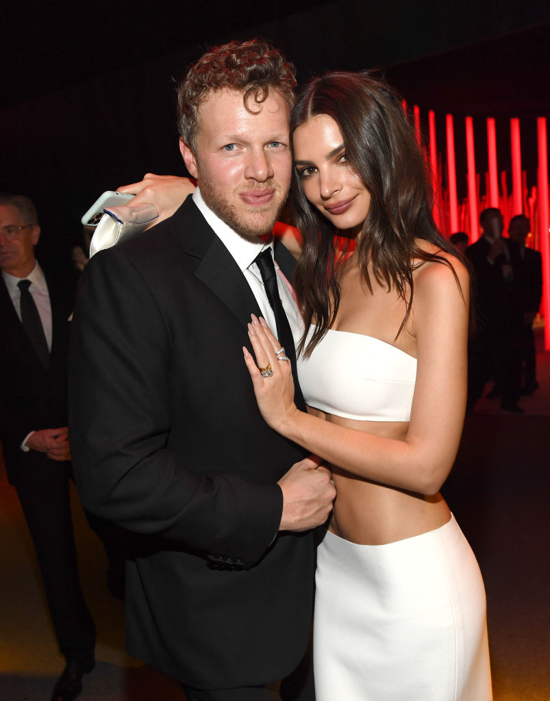 A couple poses together at an event. The woman wears a sleek, strapless white dress, while the man is in a black suit. They smile warmly