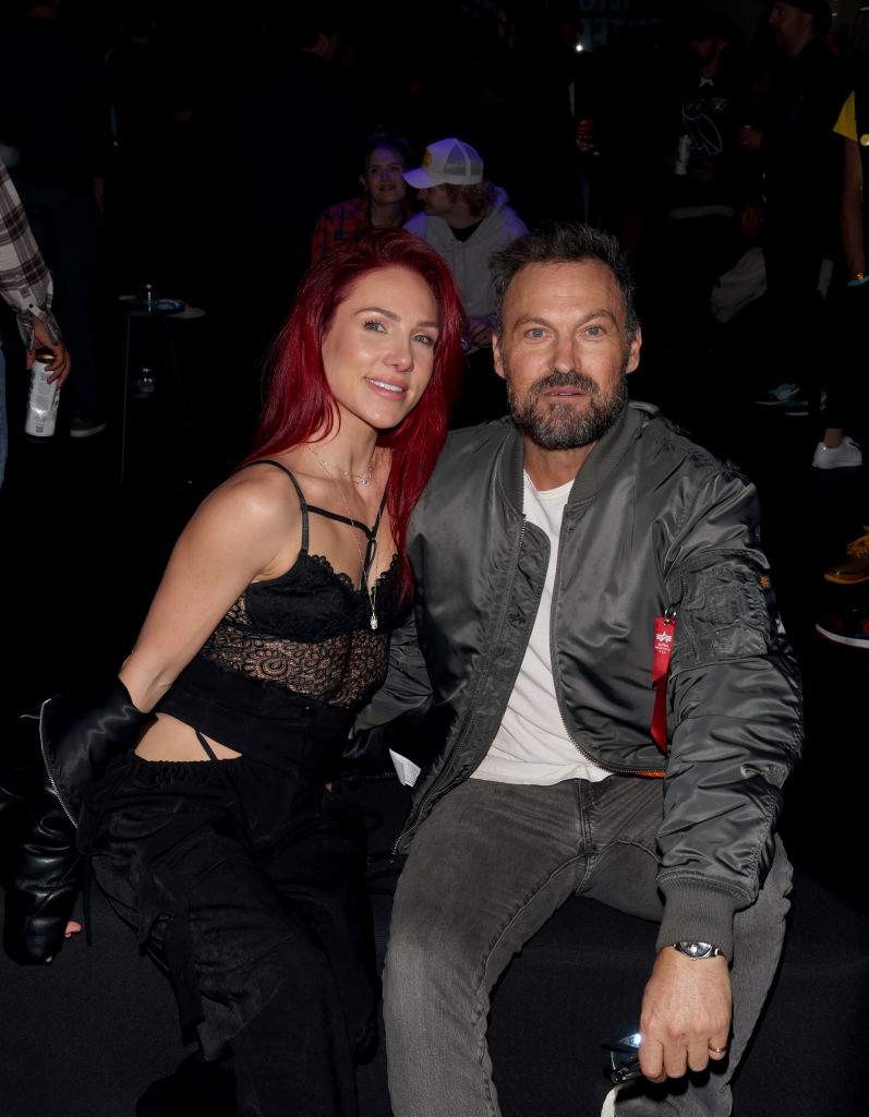 A couple sitting together at an event, the woman in a lace top and black pants, and the man in a casual gray jacket and jeans, look towards the camera