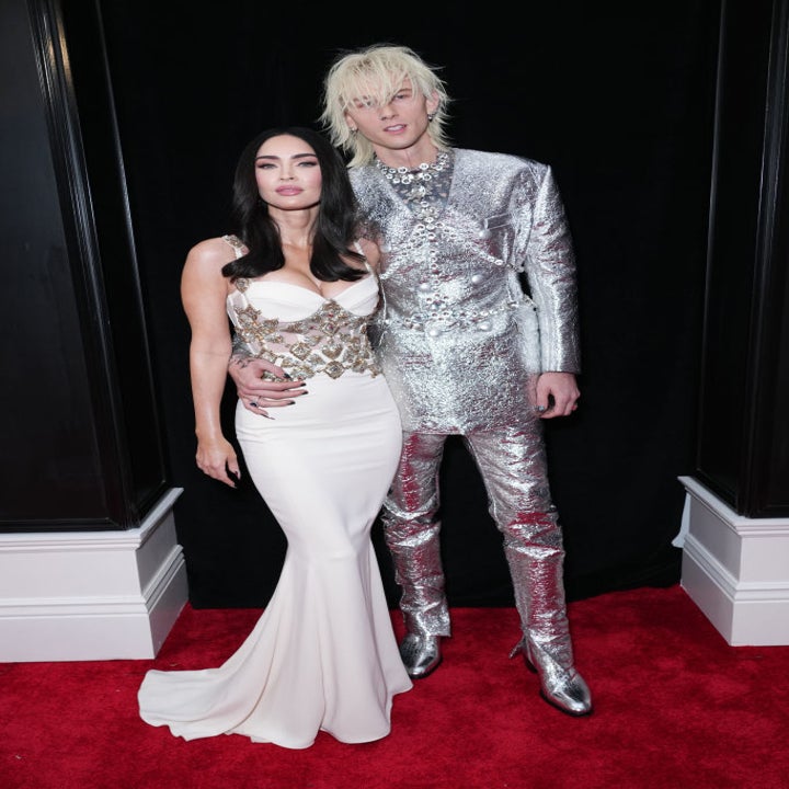 Two people on a red carpet. The woman wears a white gown with metallic details, and the man wears a shiny silver suit and boots