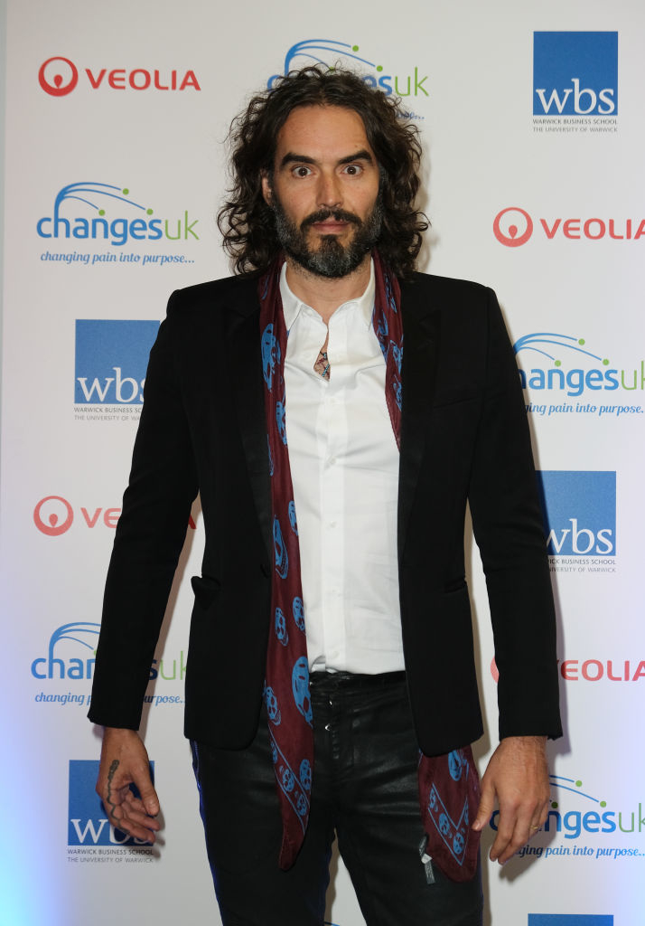 Person with long hair wearing a black jacket and patterned scarf, standing in front of a step-and-repeat with logos