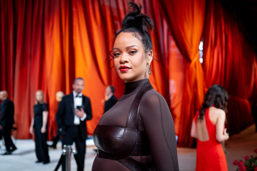 Person in elegant, semi-sheer gown poses on a red carpet with people in formal wear and draped curtains in the background