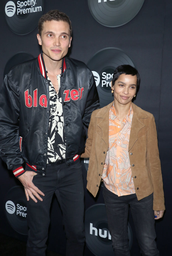 Two people stand at an event. One wears a &quot;Blazers&quot; jacket and patterned shirt, the other a patterned shirt with a suede jacket. Spotify signs in background