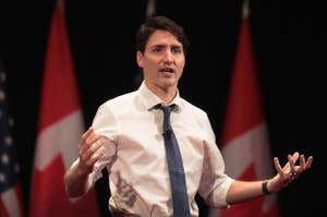 A man in a shirt and tie gestures while speaking, with flags in the background