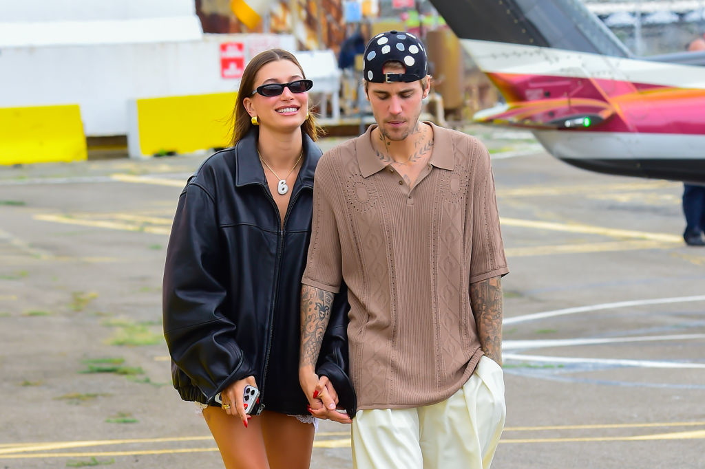 Two people walking outside, one wearing an oversized jacket and sunglasses, the other in a cable-knit shirt and cap. They are holding hands, looking relaxed