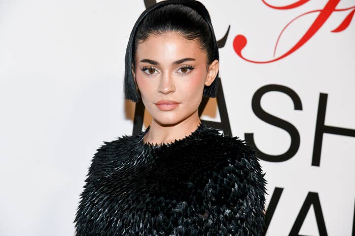Kylie, in a textured outfit, standing in front of a fashion awards backdrop. He hair is styled sleekly in a ponytail