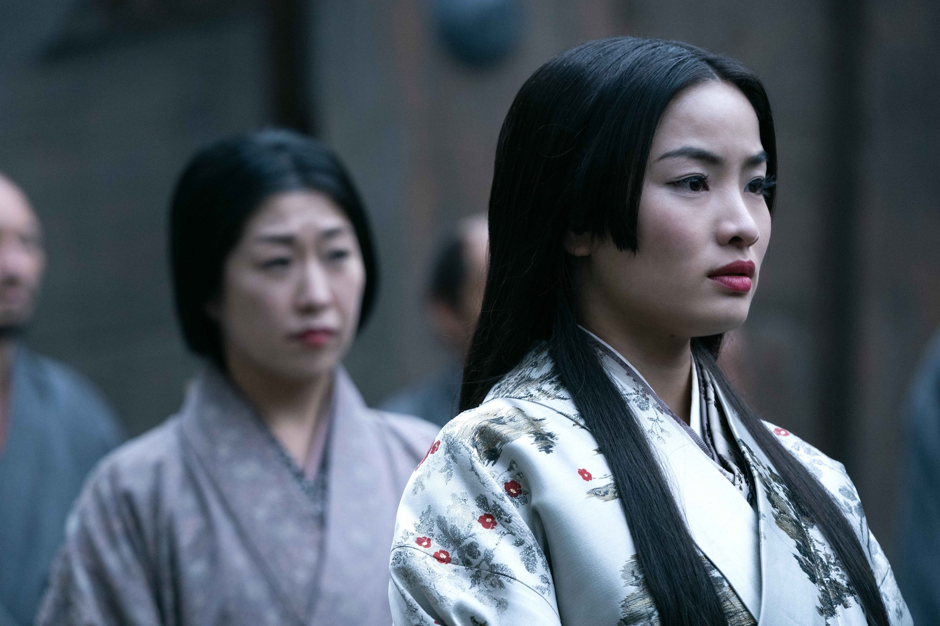 Two women in traditional Japanese kimono stand attentively, the woman in the foreground has long dark hair. It&#x27;s a scene from a TV or movie set