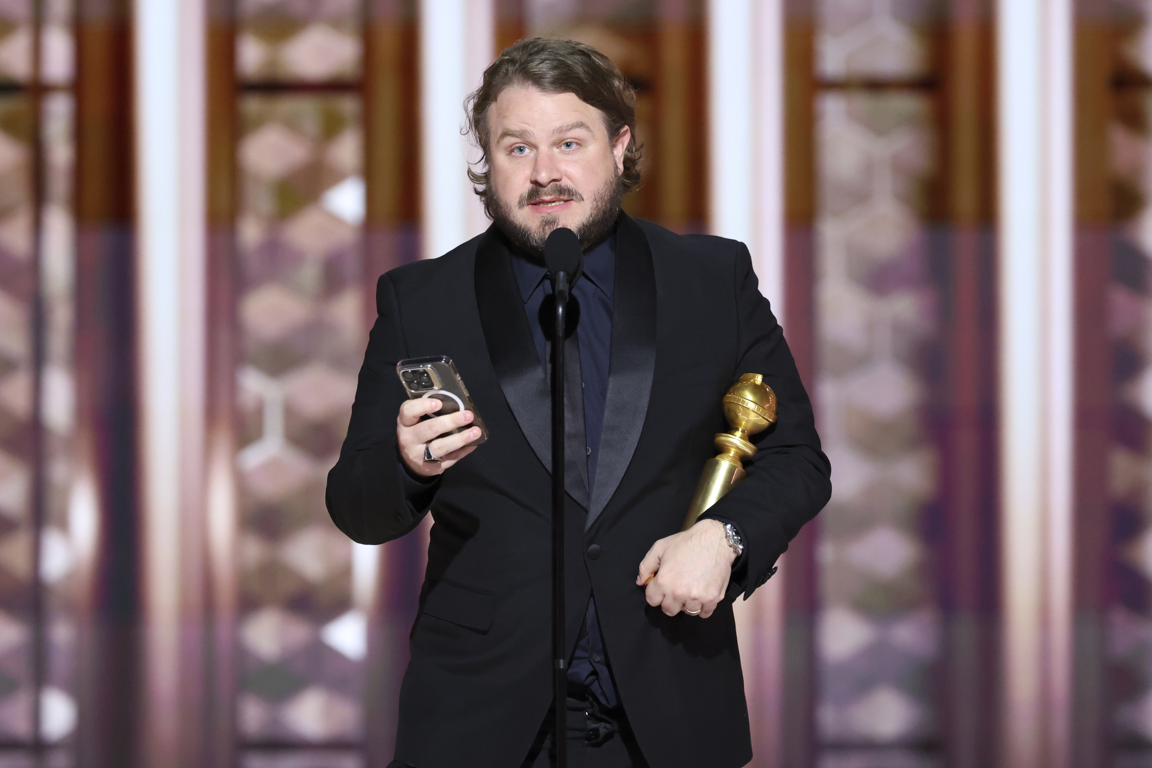 Brady Corbet in formal suit holding a golden trophy and a phone, speaking into a microphone on stage