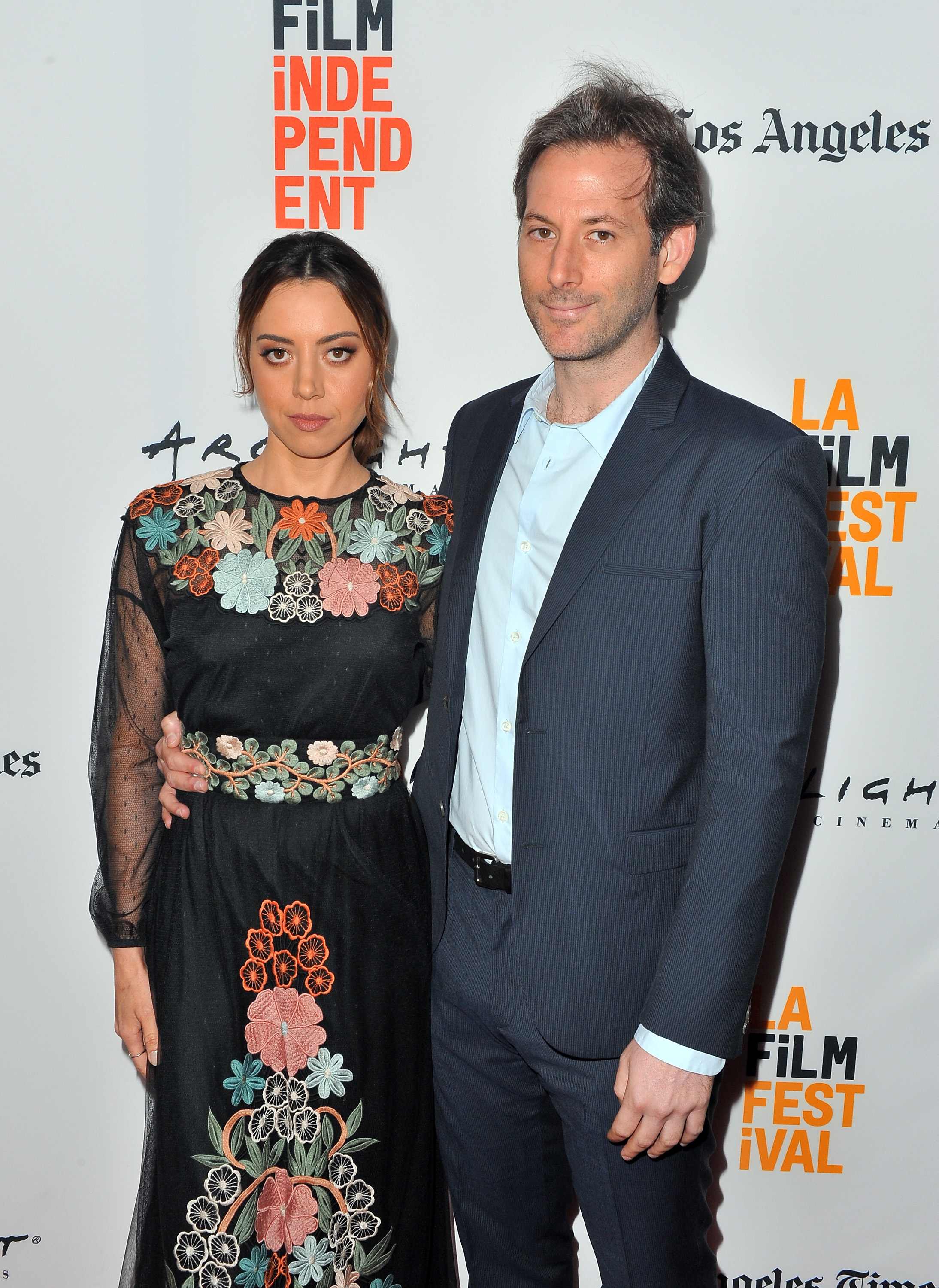 Aubrey Plaza and Jeff Baena on a red carpet; the woman wears a floral embroidered dress, the man is in a suit. The backdrop has film festival logos