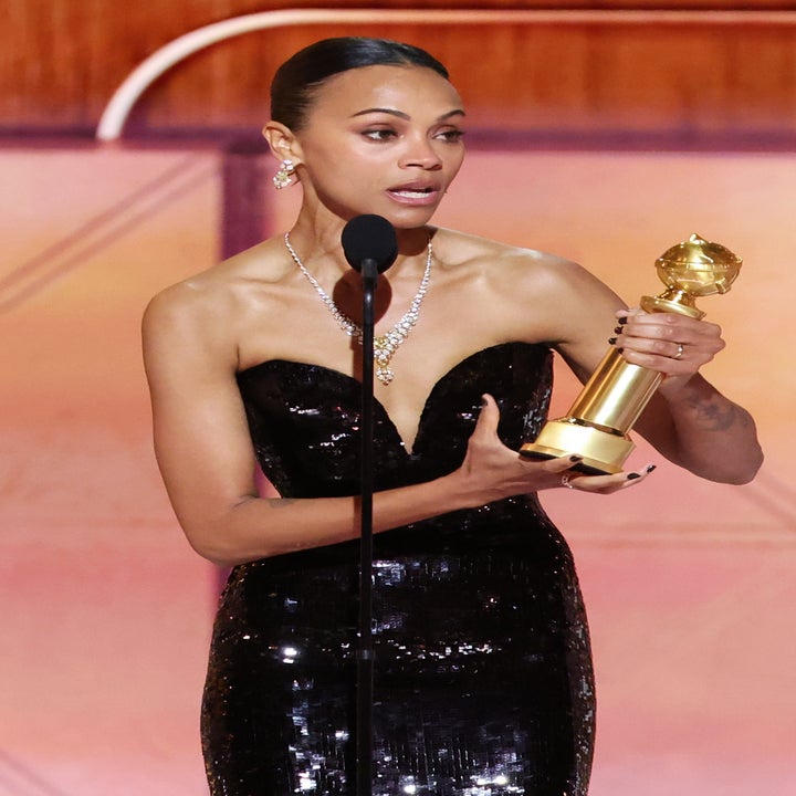 Zoe Saldaña in a strapless, sequin gown holds a golden award trophy while speaking into a microphone on stage