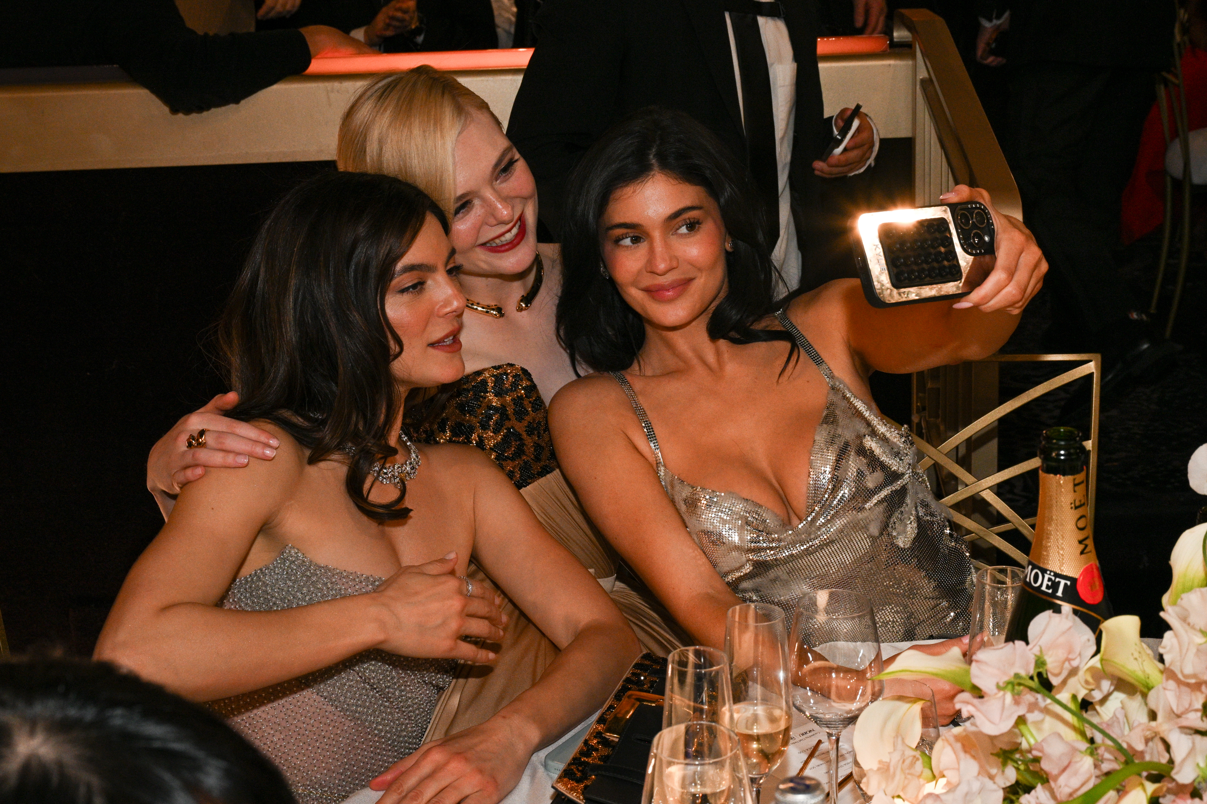 Three women in elegant evening gowns pose for a group selfie at a formal event, smiling amid champagne and floral table decor