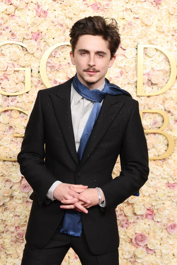 Person in a black suit and a blue textured scarf stands in front of a floral backdrop