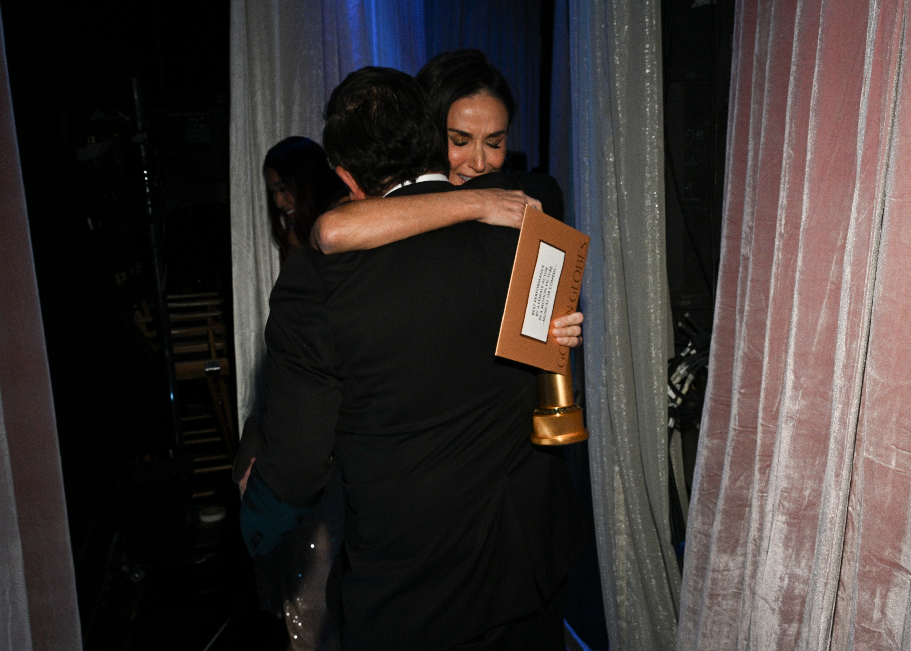 Person embraces an award recipient holding a trophy behind a curtain. Both are wearing formal attire