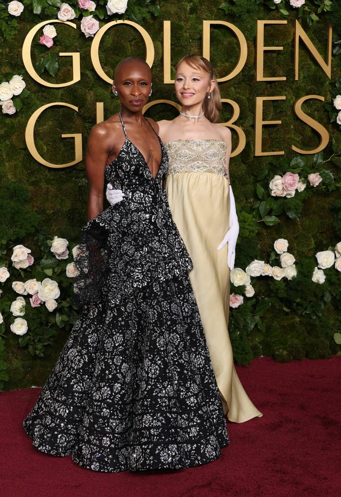 Cynthia Erivo and Ariana Grande on a red carpet at the Golden Globes, one in a black halter gown with floral patterns, and the other in a strapless yellow gown with white gloves