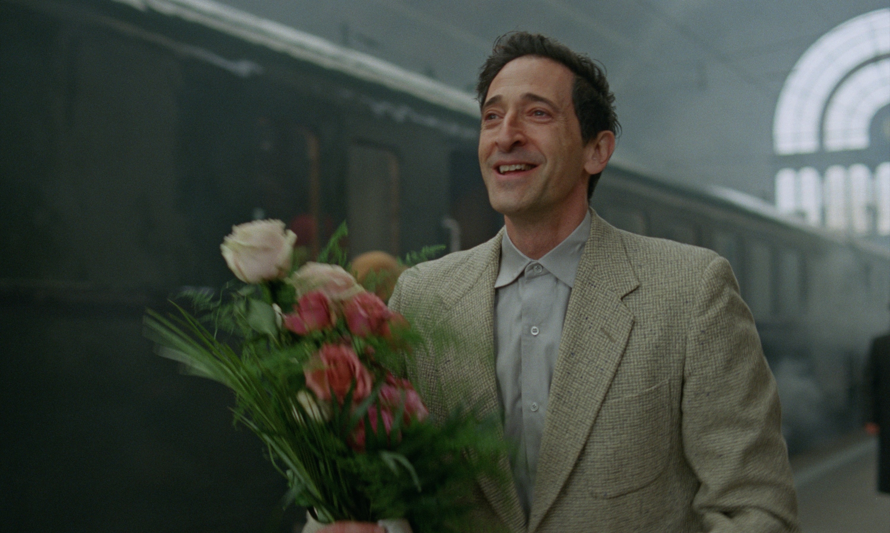 A person smiling and holding a bouquet stands in front of a vintage train at a station. They&#x27;re wearing a textured suit jacket and light-colored shirt