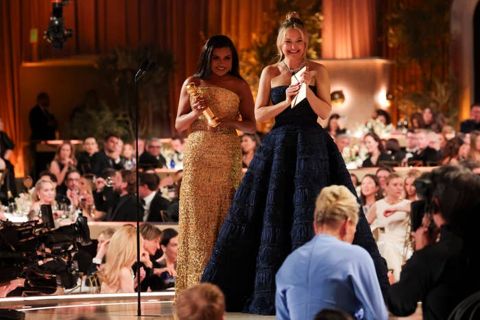 Two women stand on stage at an awards event. One in a shimmering gown holds a trophy, while the other wears a textured, elegant gown and smiles
