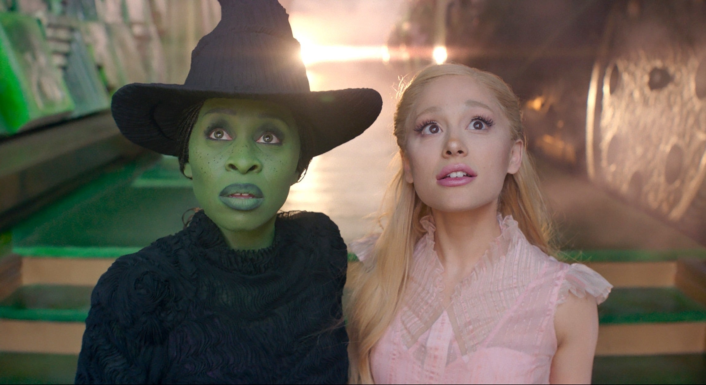 Two actors in a fantasy setting; one wearing a witch&#x27;s hat and dark clothing, the other in a light, sleeveless dress, both looking upward