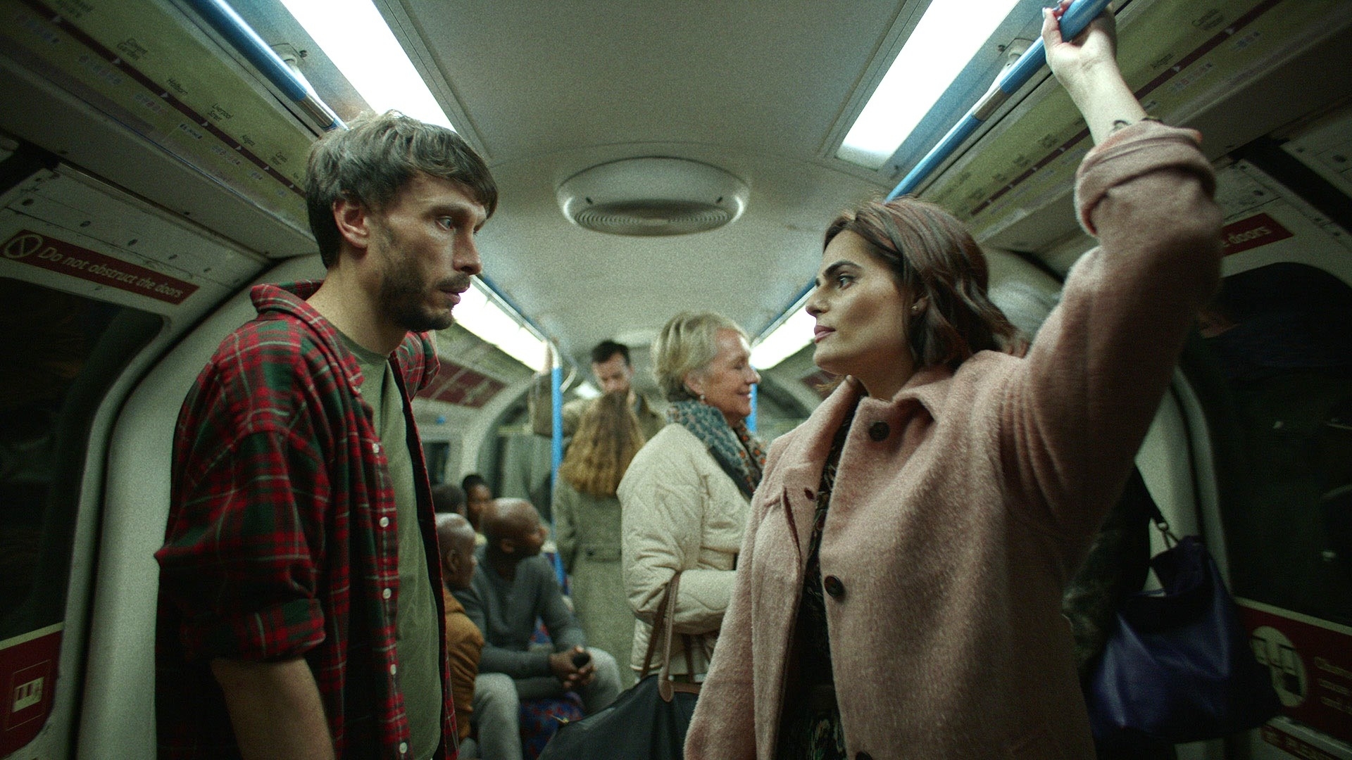 Two people stand facing each other on a crowded subway train, engaged in a tense conversation