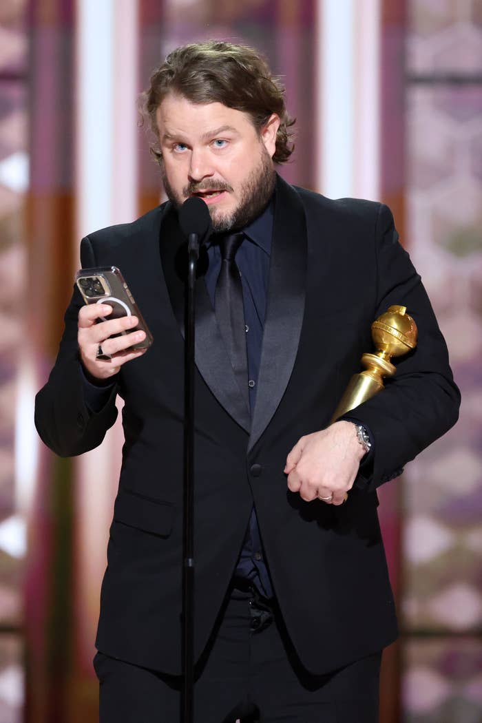 Brady Corbet in a suit holding an award and phone, speaking into a microphone