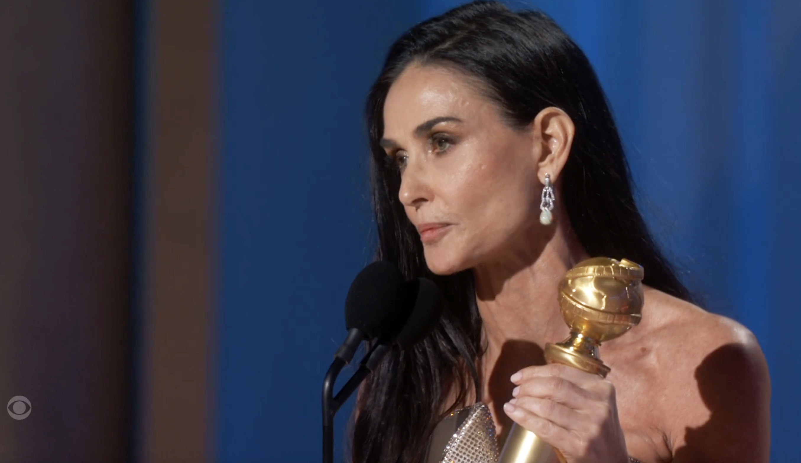 A person in a strapless gown holds a Golden Globe award, speaking into a microphone on stage