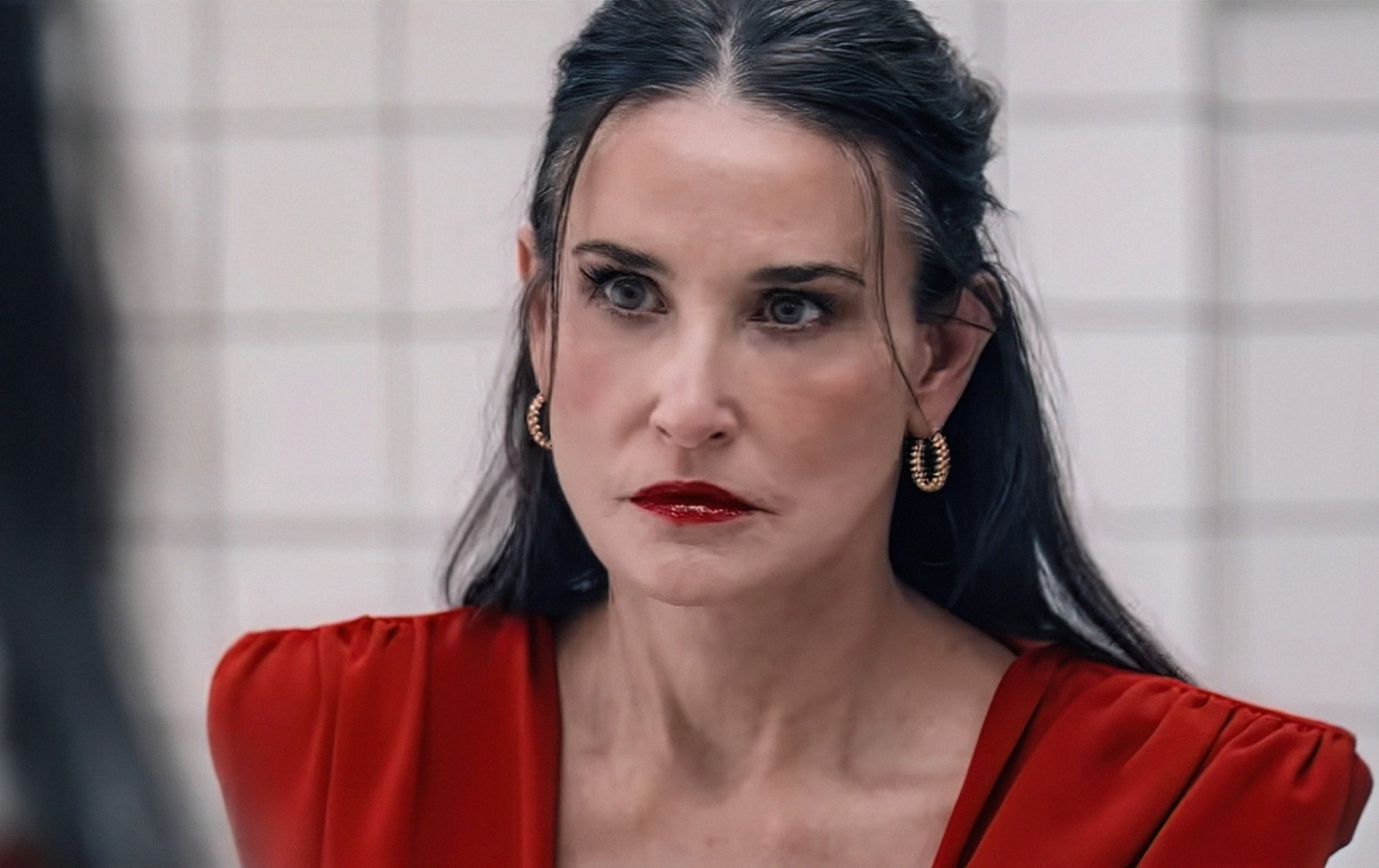 Person with long hair and hoop earrings in a serious expression, looking in the mirror, wearing an elegant outfit in a bathroom setting