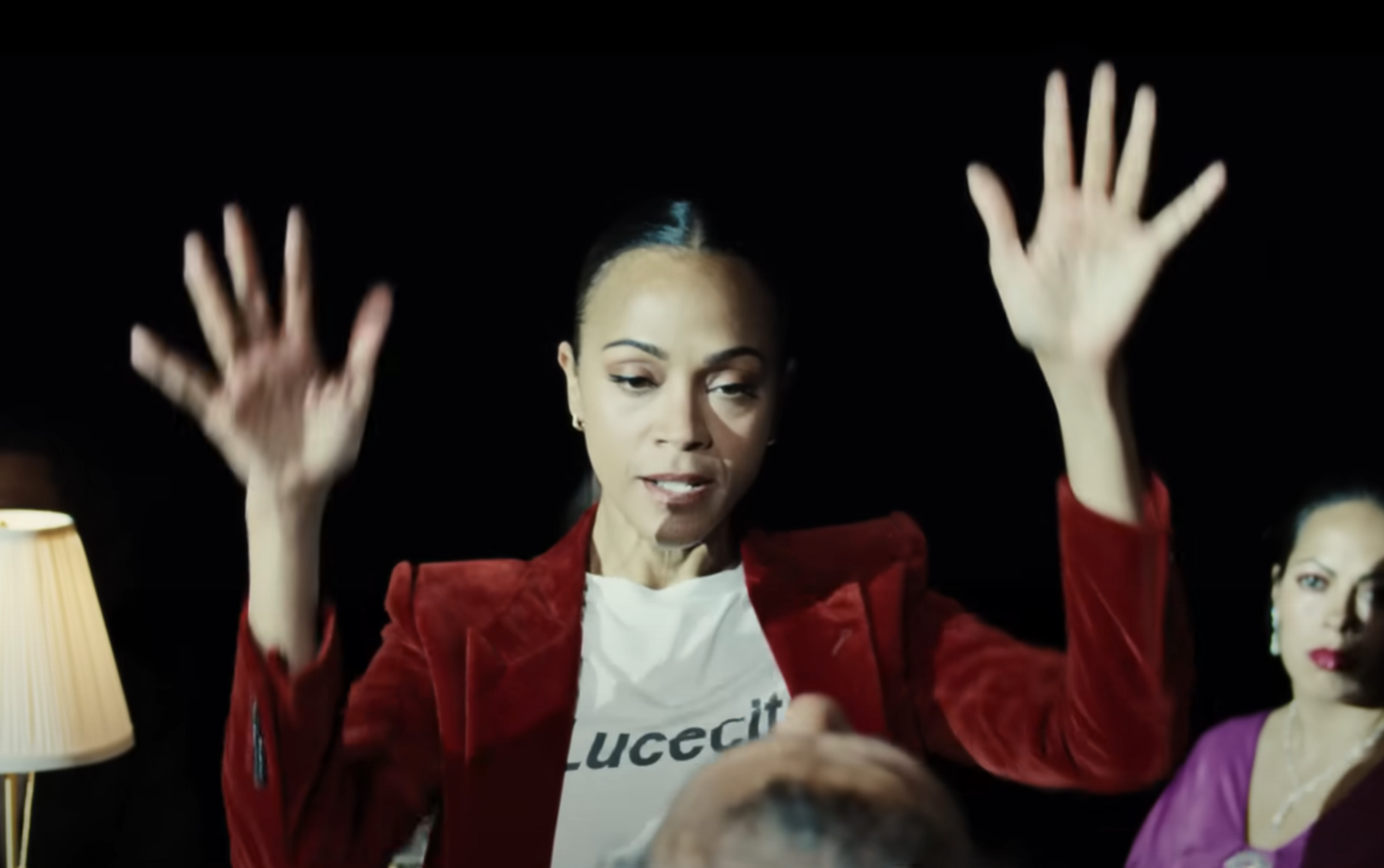 Person in a velvet blazer and graphic tee raises hands dramatically, with another person watching intently in a dimly lit setting