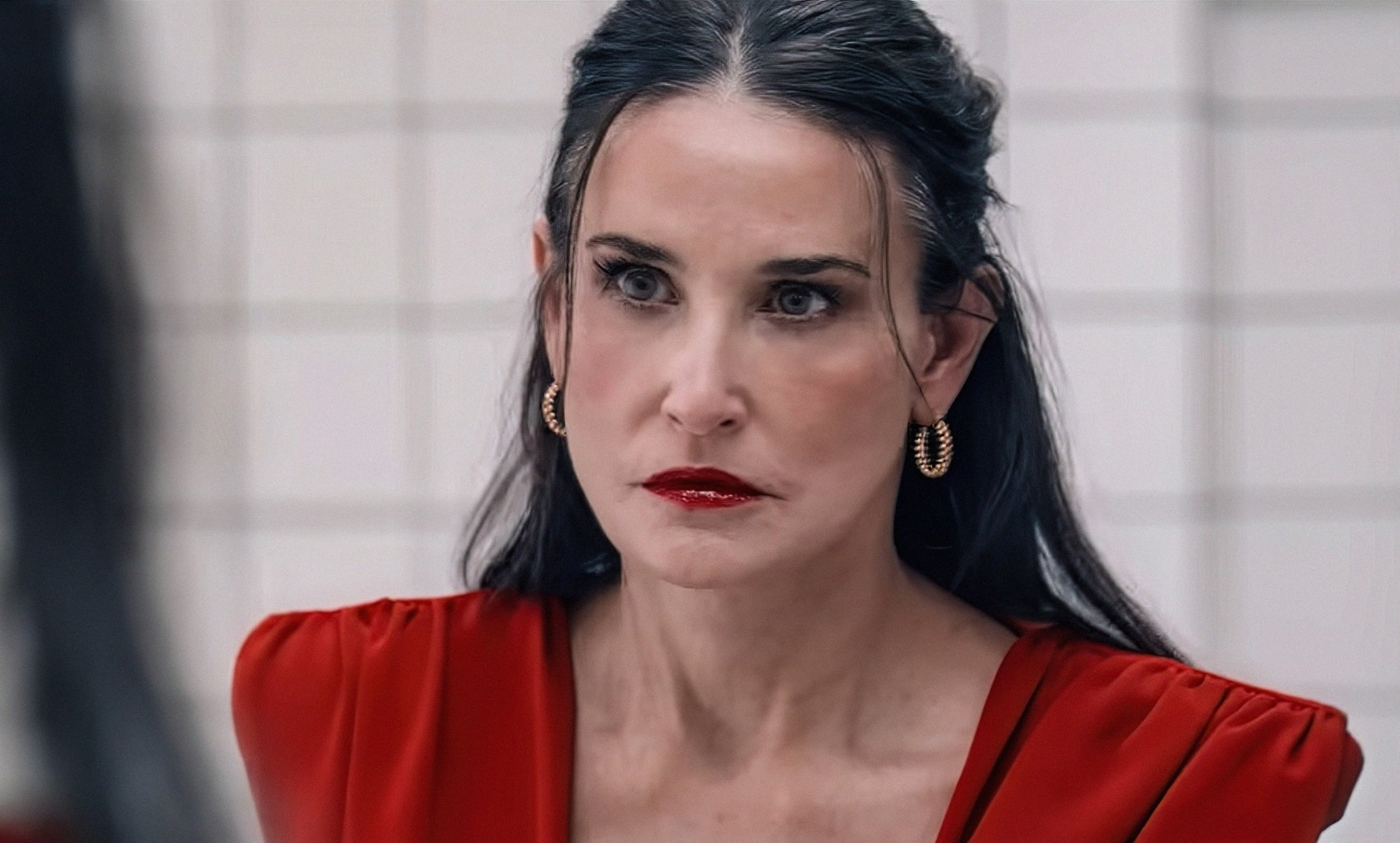 Person with long dark hair and gold earrings wears a stylish outfit, looking intensely into the mirror in a white-tiled room