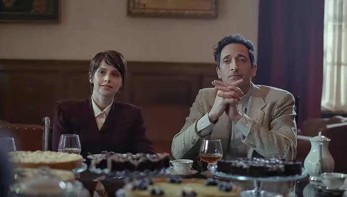 Two people sit at a table with desserts and drinks in front of them, appearing to be in a formal setting