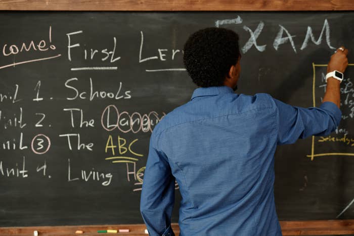 Person writing on a blackboard with headings &quot;First Lesson&quot; and &quot;EXAM,&quot; and a list of units like &quot;Schools&quot; and &quot;Living.&quot;