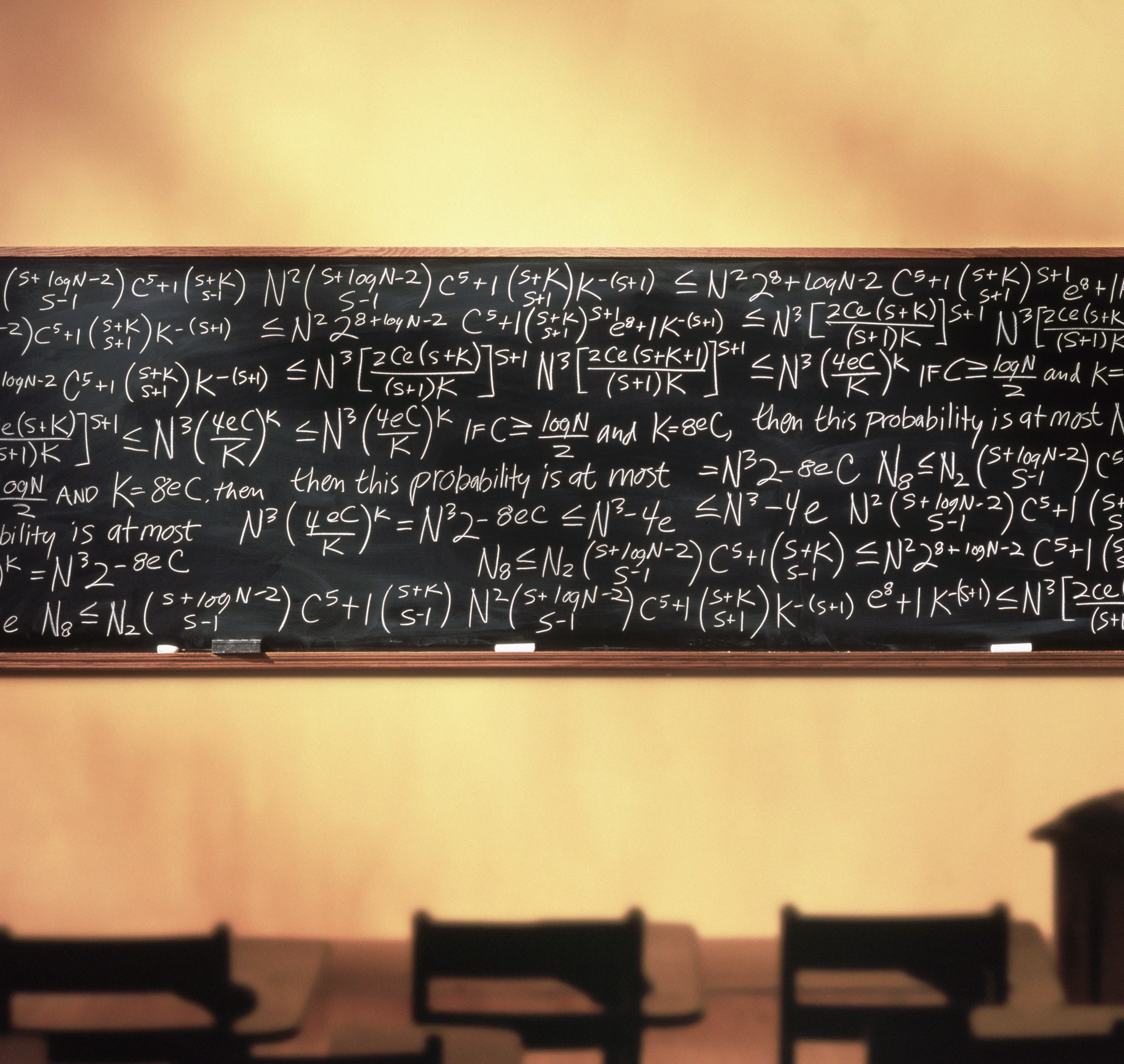 A classroom chalkboard covered with complex mathematical equations and formulas, with empty desks in front