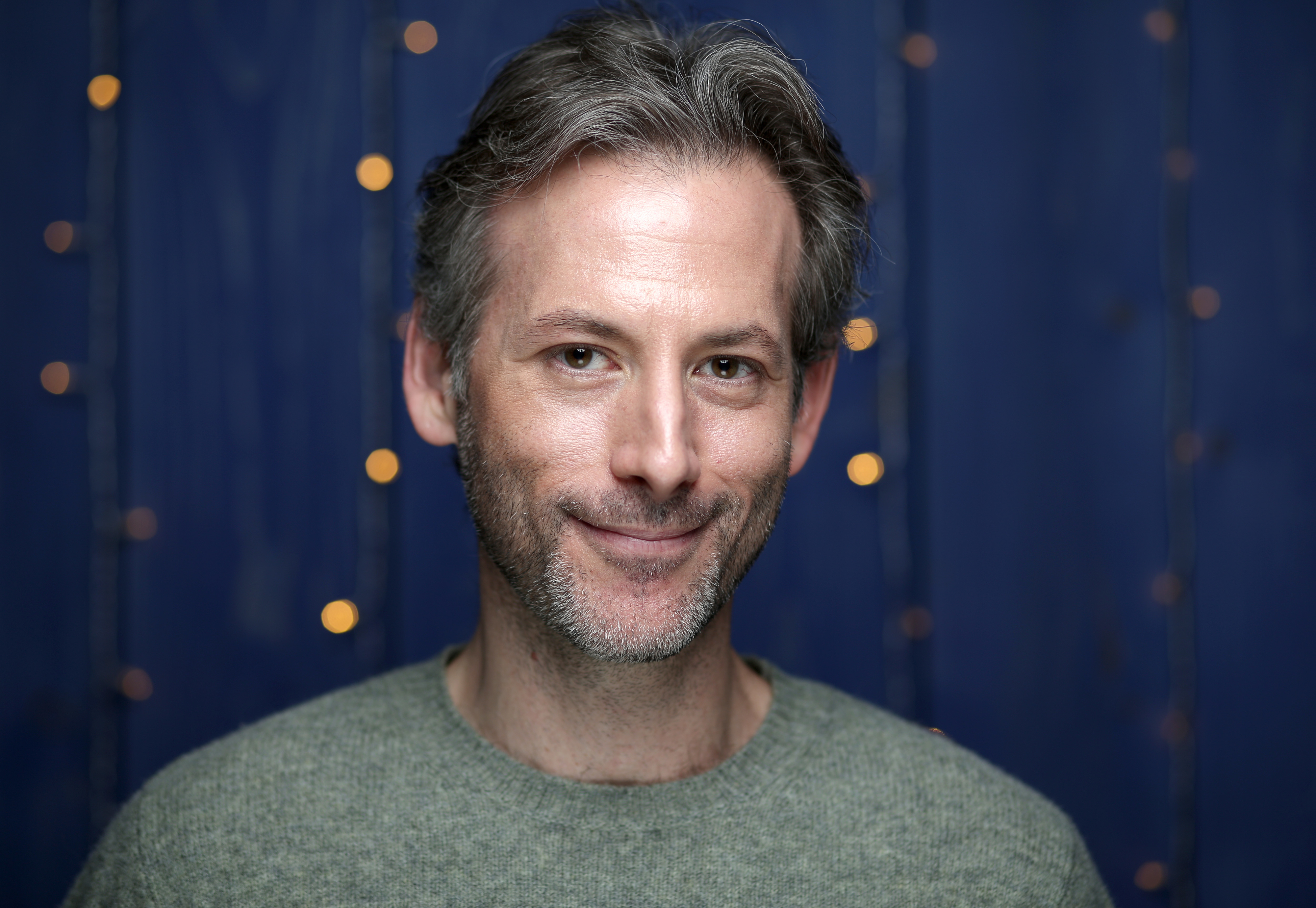 Jeff Baena with short hair and light facial hair, smiling slightly, against a blurred background with lights