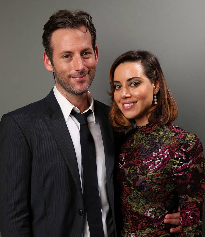 Jeff Baena in a suit and tie stands next to Aubrey Plaza wearing a floral-patterned high-neck dress, both smiling at the camera against a neutral background