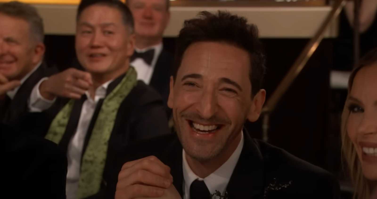 Adrien Brody in a formal attire smiling and holding hands at a formal event. People are visible in the background, also dressed formally