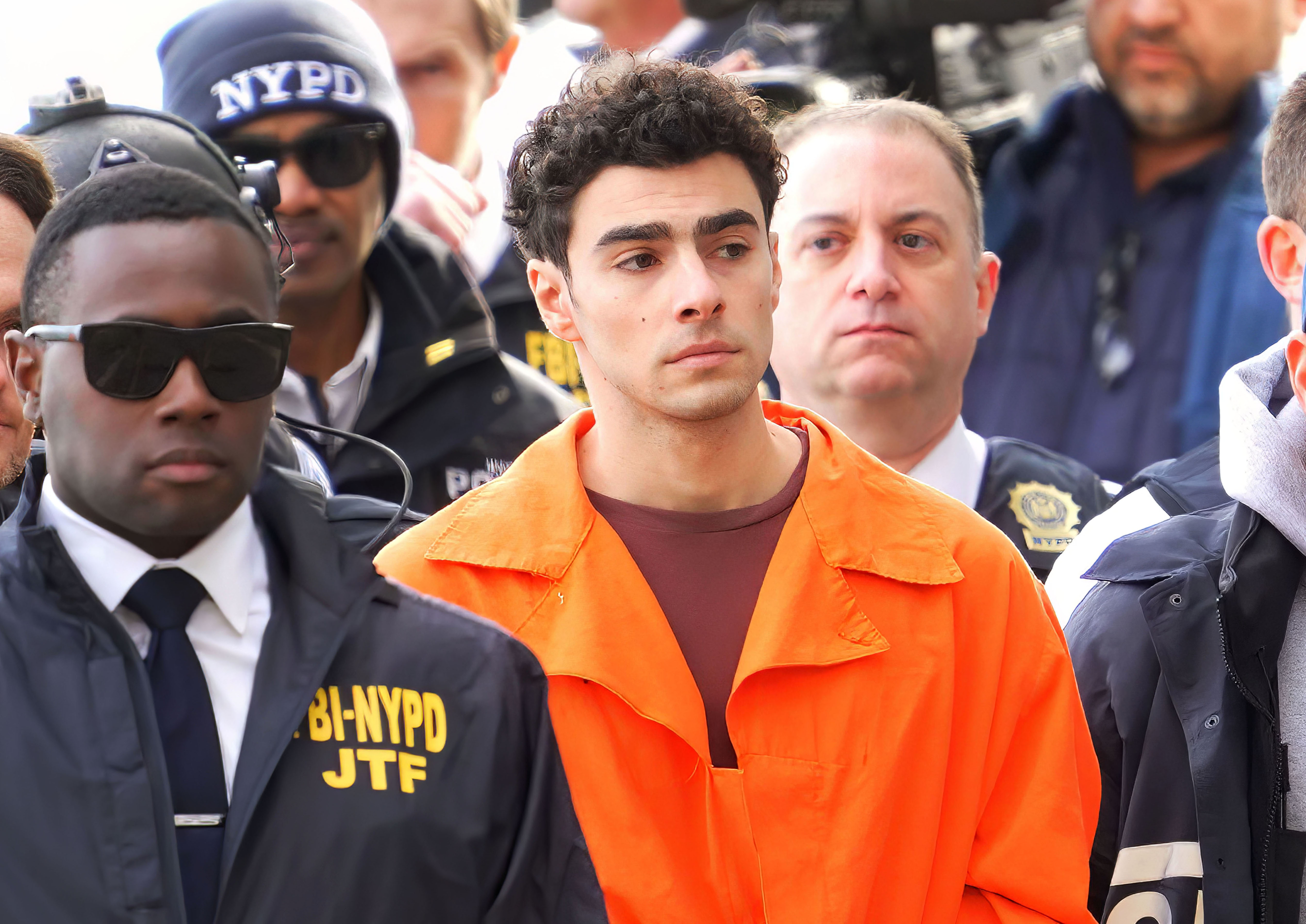 A person in an orange jumpsuit, surrounded by law enforcement officers, including one in a jacket with FBI-NYPD JTF insignia