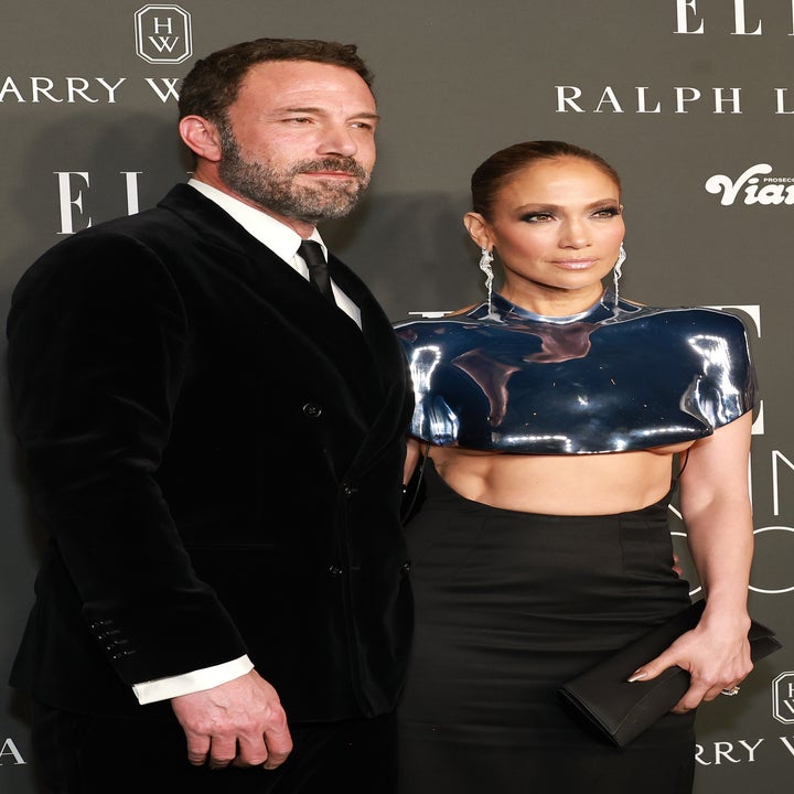 Two individuals pose together at a formal event; one in a velvet suit, the other in a metallic-themed top and sleek skirt, holding a clutch