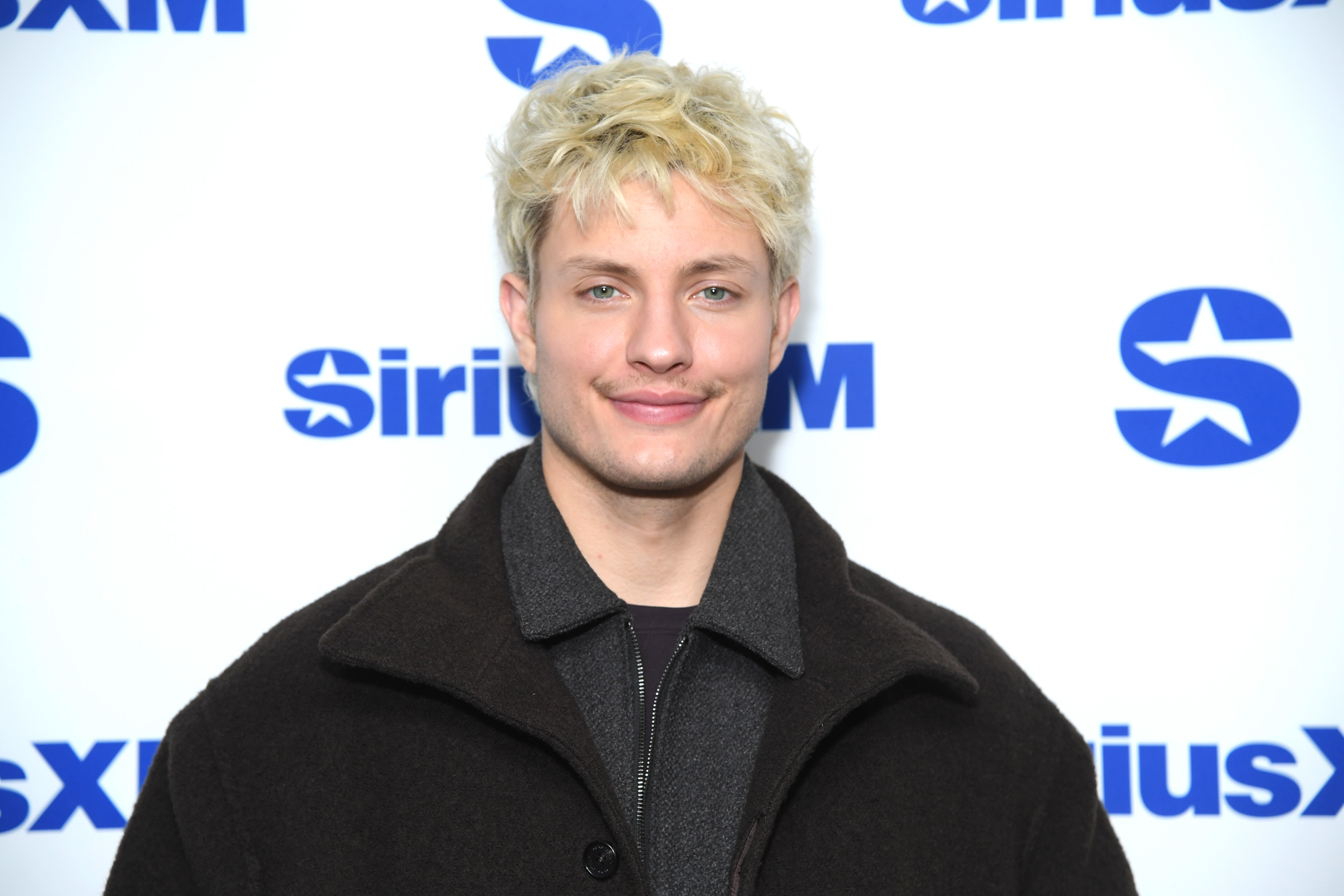 Matt Rife smiling at a SiriusXM event, wearing a dark coat over a shirt