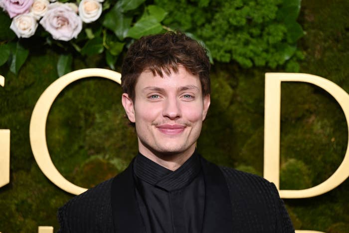 Matt Rife smiling in front of a backdrop with flowers and large letters. Wearing a dark textured suit jacket and shirt