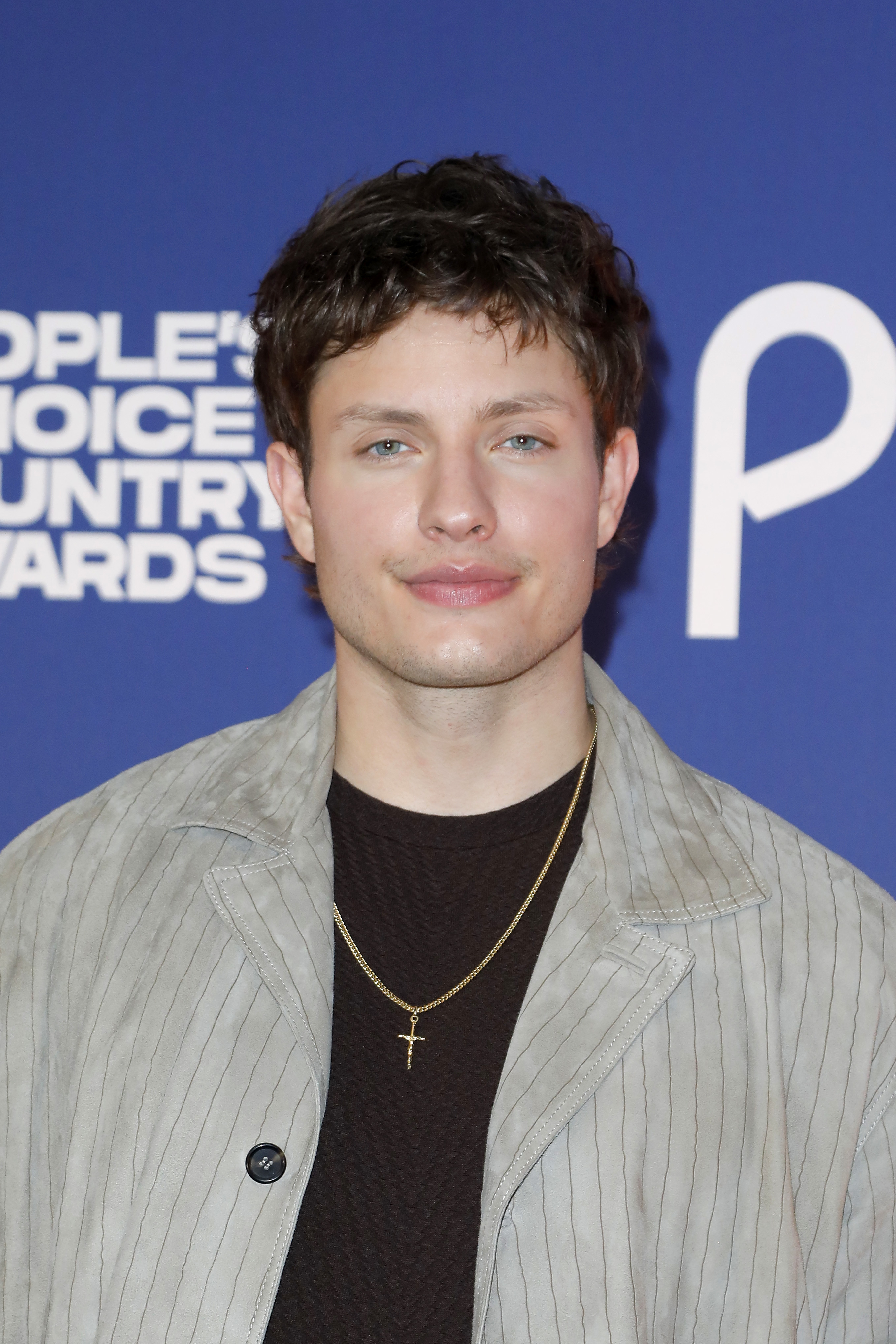 Matt Rife on red carpet in a striped jacket and dark shirt, wearing a necklace with a cross pendant, attending an event with &quot;People&#x27;s Choice&quot; backdrop