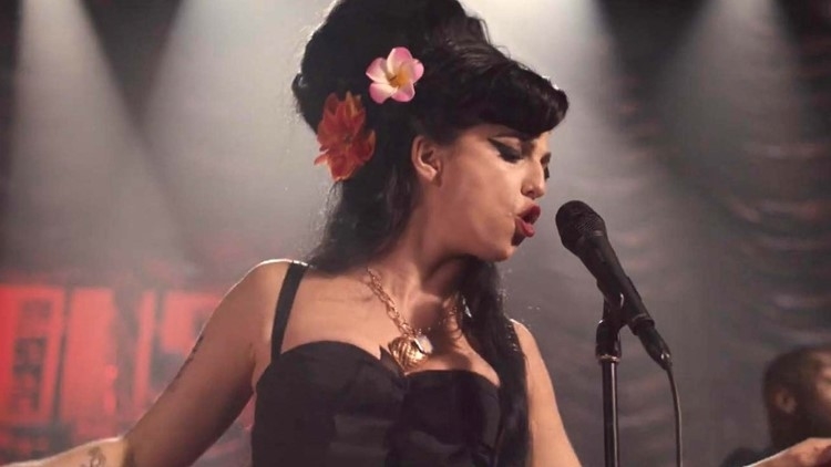 Singer performing on stage, wearing a vintage-style dress with flower accents in her hair, passionately singing into a microphone