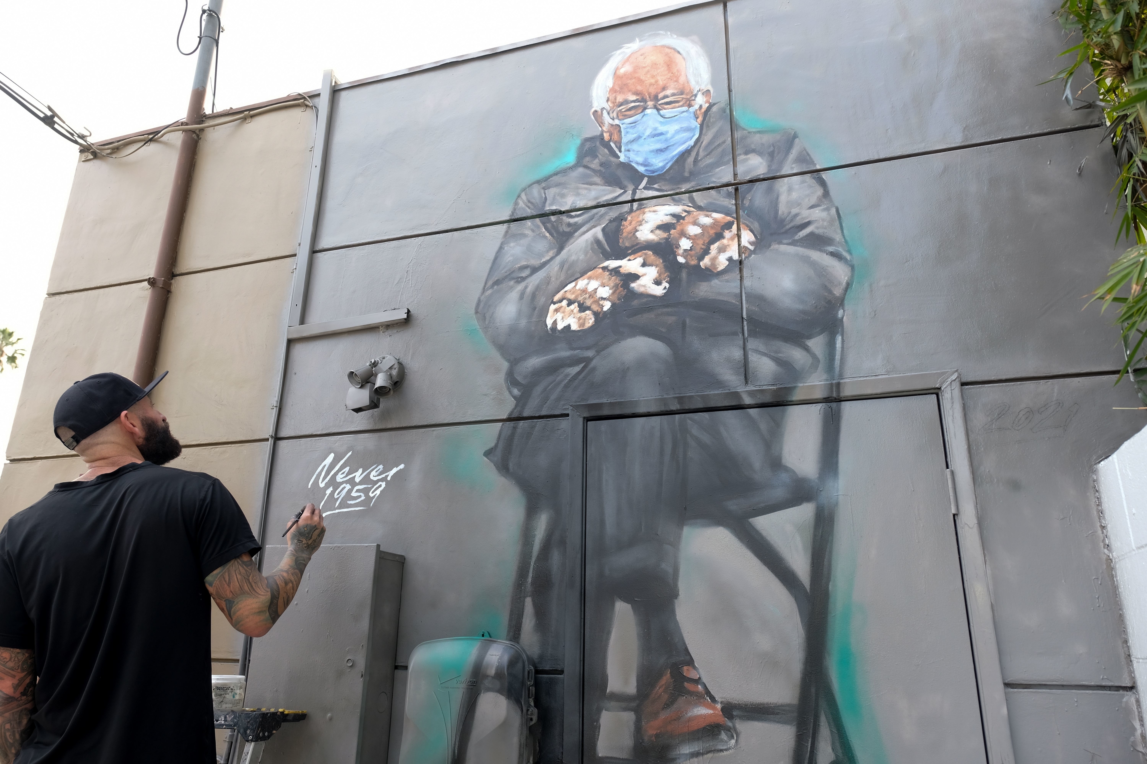 A mural depicts a Sanders sitting with crossed legs, wearing a mask, mittens, and a winter coat. An artist is working on the mural