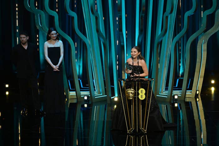 Three people stand on a stage, one giving a speech at an awards ceremony. Elegant formal attire is worn by the group