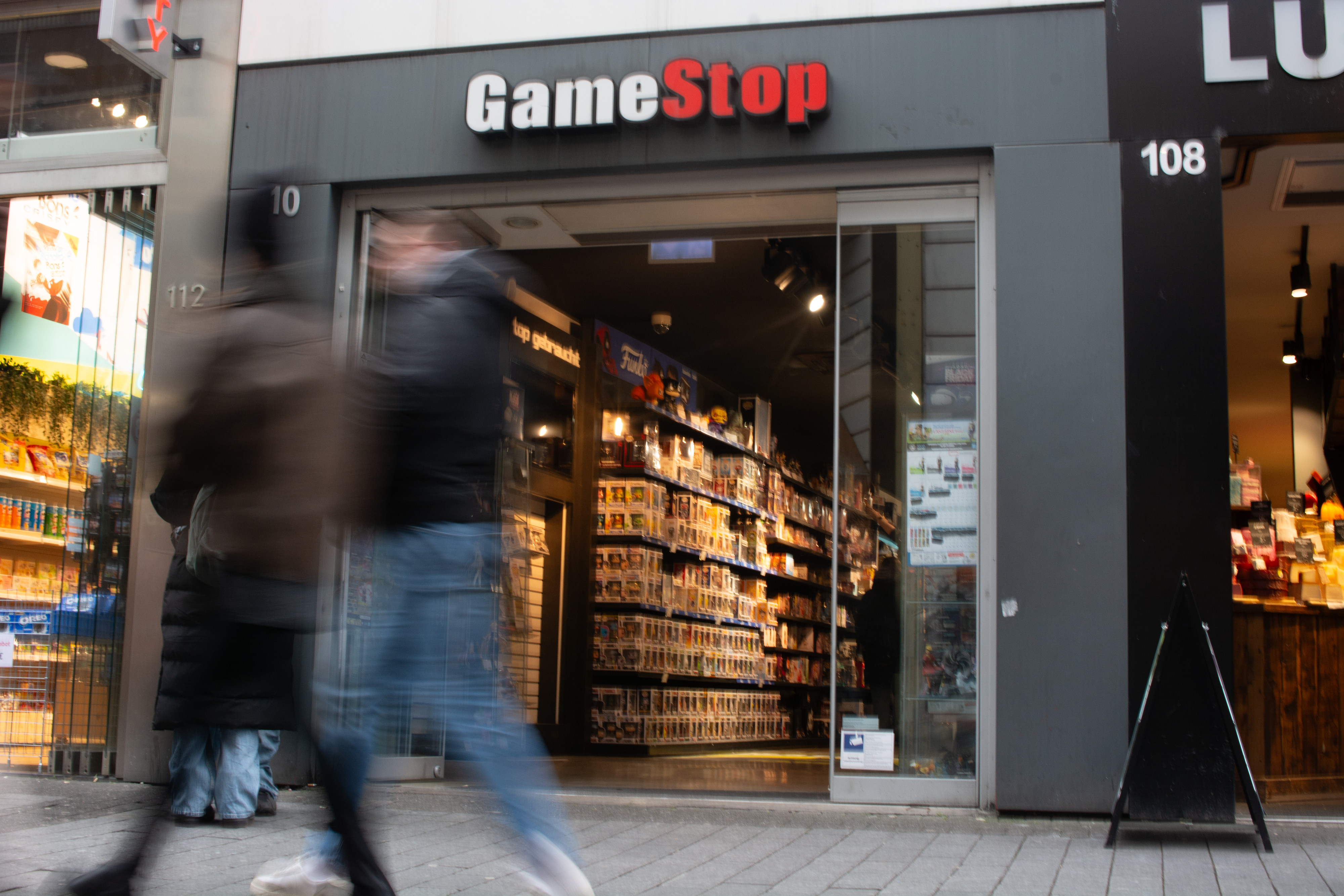 GameStop storefront with blurred pedestrians walking by, shelves visible inside through the glass doors